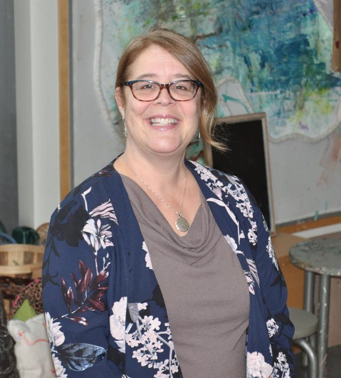 Dr. Jessica Brian is wearing a grey shirt and a blue camisole with a white floral pattern. She is smiling for a photograph looking at the camera.